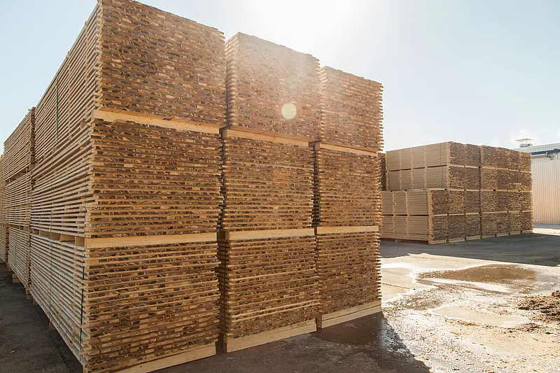 Stacks of cut and dried wooden planks arranged in large piles at a lumber yard, with sunlight shining from above