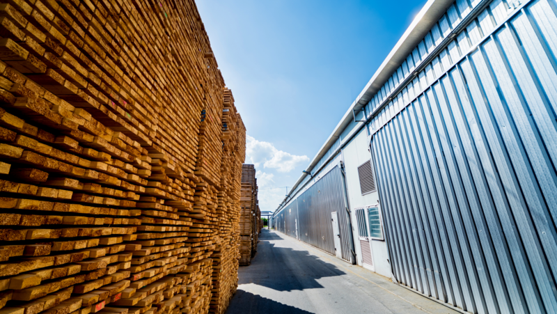 Stacks of kiln-dried timber neatly piled beside a modern industrial drying facility under a clear blue sky.