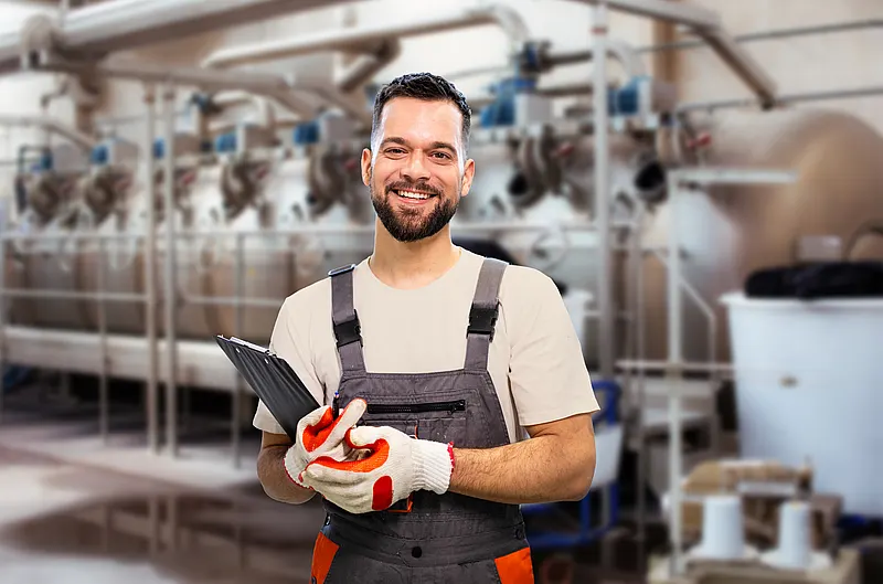  纺织品干燥厂内，佩戴手套、手持夹板的微笑工人，背景为大型干燥设备。