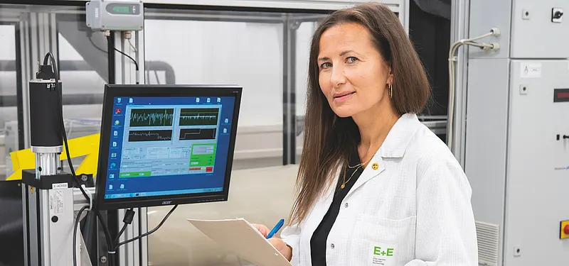 In a calibration laboratory, a woman with long hair reviews data on a monitor while taking notes on a clipboard.